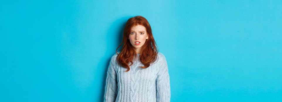 Confused redhead girl in sweater staring at camera, raising eyebrow and feeling puzzled, standing over blue background.