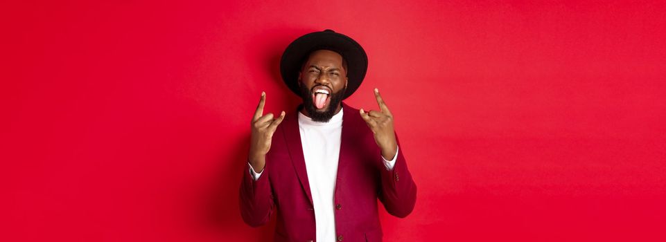 Happy african american man having fun on party, showing tongue and rock-n-roll gesture, celebrating New Year, standing against red background.