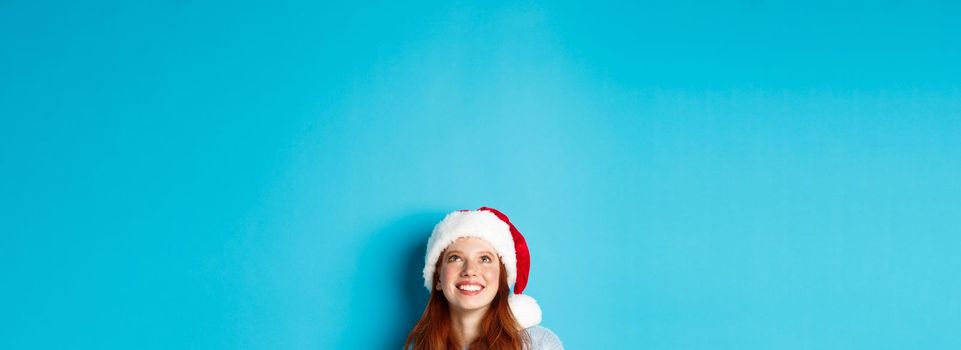 Winter holidays and Christmas eve concept. Head of cute redhead girl in santa hat, appear from bottom and looking up at copy space, staring logo, standing over blue background.