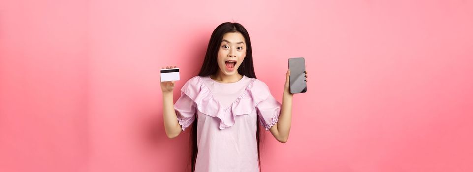 Online shopping. Excited asian woman showing plastic credit card with empty smartphone screen, advertising internet shop, standing on pink background.