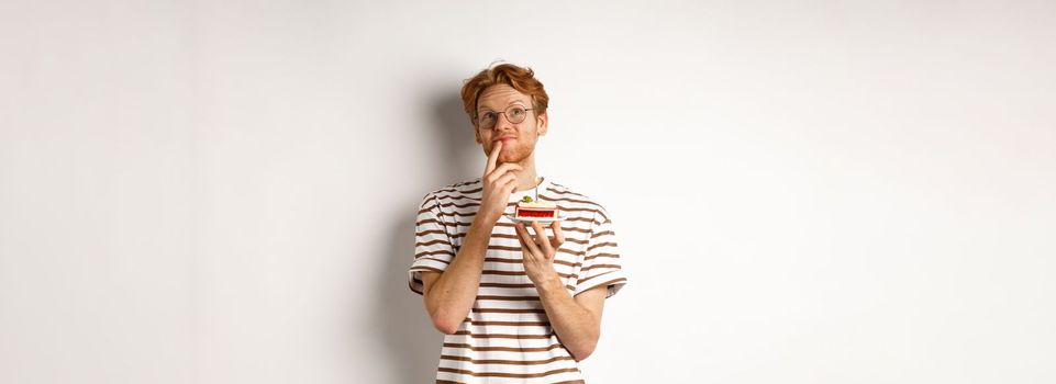 Holidays and celebration concept. Happy young man with red hair and glasses having birthday, holding cake with candle and thinking of b-day wish, standing over white background.