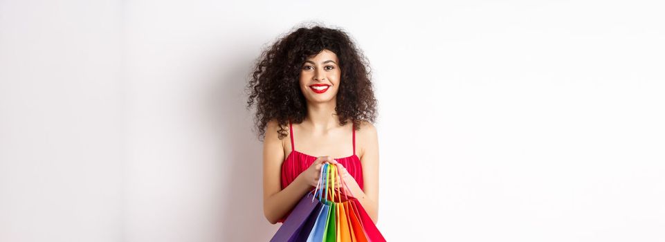 Image of stylish caucasian woman in red dress and makeup, holding shopping bags and smiling, standing over white background.