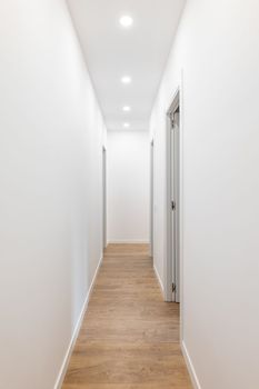 Long corridor with white smooth walls, bright light pours from spotlights from the ceiling, wooden laminate flooring, open doors on both sides to separate living rooms