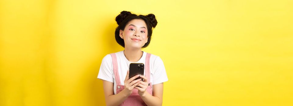 Cute stylish asian girl using mobile phone, wearing glamour pink makeup and summer clothes, standing on yellow background.