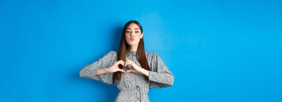 Valentines day. Romantic girl pucker lips for kiss, showing heart gesture, say I love you and look at camera, standing in dress on blue background.