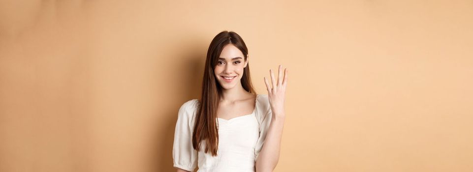 Attractive young woman show fingers number four, smiling and looking confident, standing on beige background.