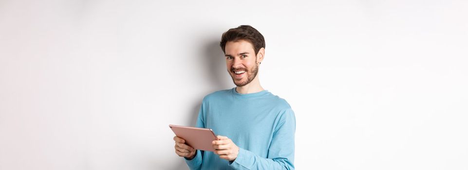 E-commerce and technology concept. Young caucasian man smiling at camera, holding digital tablet, standing over white background.