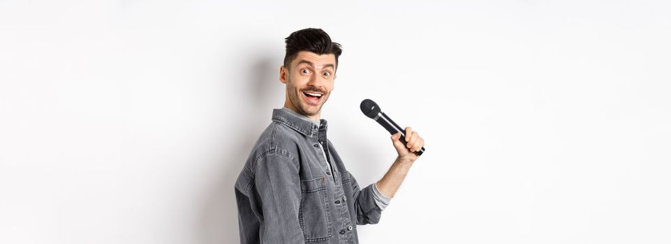 Profile of handsome smiling man holding mic, turn head at camera with excited face, singing karaoke and perform standup, standing on white background.