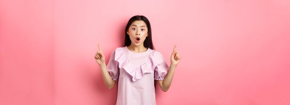 Excited asian teen girl pointing fingers up, saying wow and showing promo deal, standing in dress against pink background.