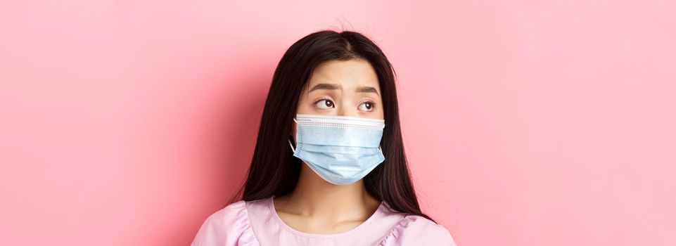 Covid-19, pandemic and quarantine concept. Close-up of sad asian girl in medical mask feeling lonely during coronavirus, standing against pink background.