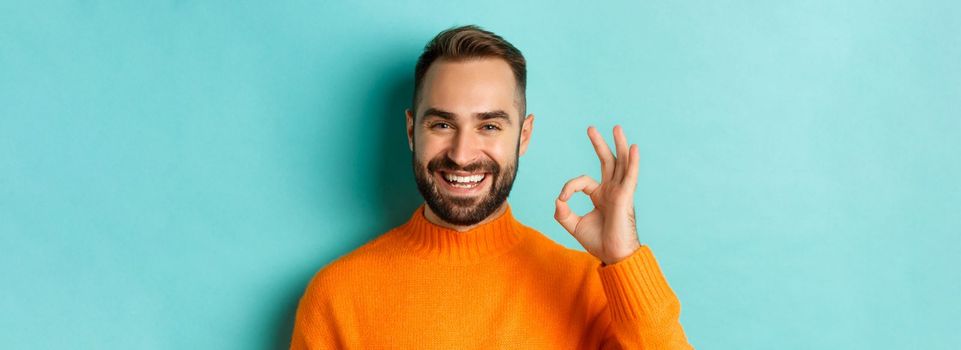 Close-up of assertive young man assuring everything ok, showing okay sign and smiling, yes or positive answer, standing over light blue background.