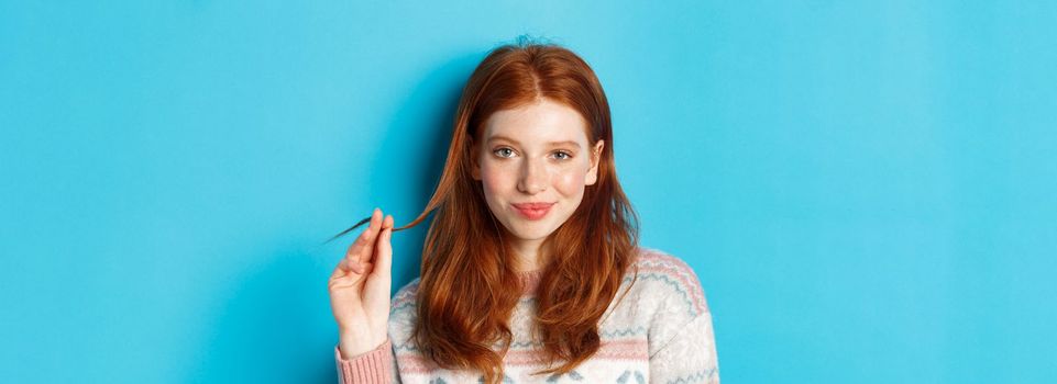 Close-up of confident and sassy redhead teen girl looking at camera pleased, playing with hair strand and smirking, standing over blue background.