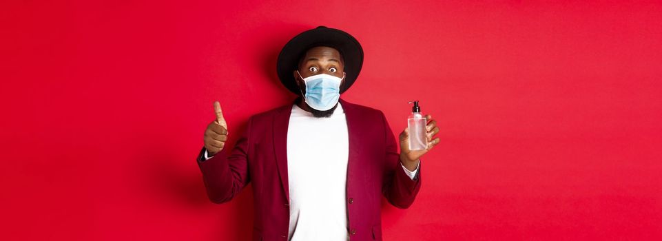 Covid-19, quarantine and holidays concept. Impressed black man in party outfit and medical mask, showing thumb up and hand sanitizer, standing over red background.