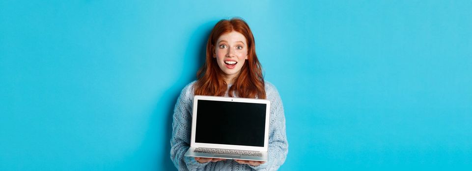 Excited redhead female freelancer showing laptop screen, staring at camera amazed, standing with computer against blue background.