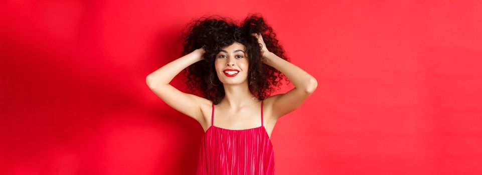 Carefree lady in red dress, touching her curly hair and smiling happy, standing on studio background. Copy space