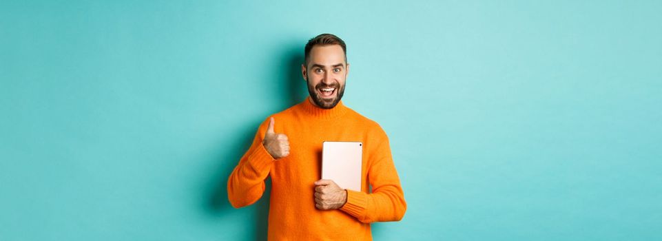 Work from home, technology concept. Handsome man holding laptop, showing thumb up, approve and like something, standing over turquoise background.