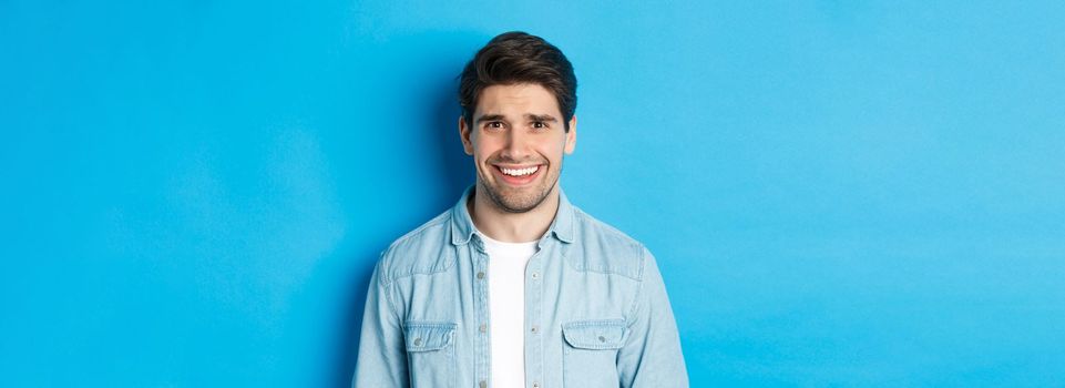 Close-up of young man feeling awkward, smile and cringe from uncomfortable situation, standing over blue background.