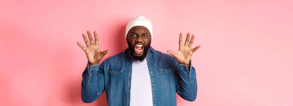 Angry african-american guy in beanie, scare you, roaring and screaming, showing hands, standing ove rpink background.