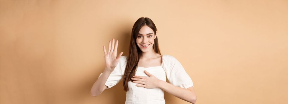 Cute smiling girl make promise, put hand on heart and tell truth, being honest, swearing to you, standing on beige background.