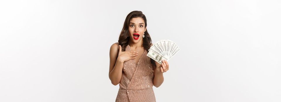 Shopping concept. Elegant woman in glamour dress holding money and looking surprised. winning prize, standing over white background.