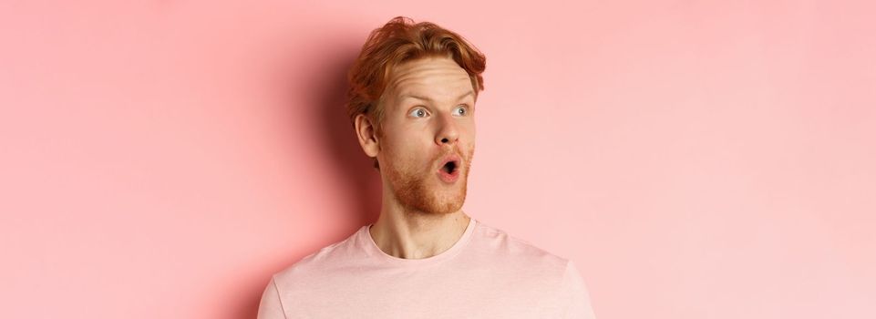 Close up of shocked redhead man with beard, saying wow, looking left with amazed face, standing over pink background.