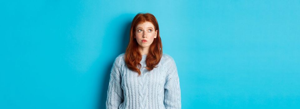 Dreamy redhead girl thinking or making decision, looking at upper left corner logo, standing against blue background.