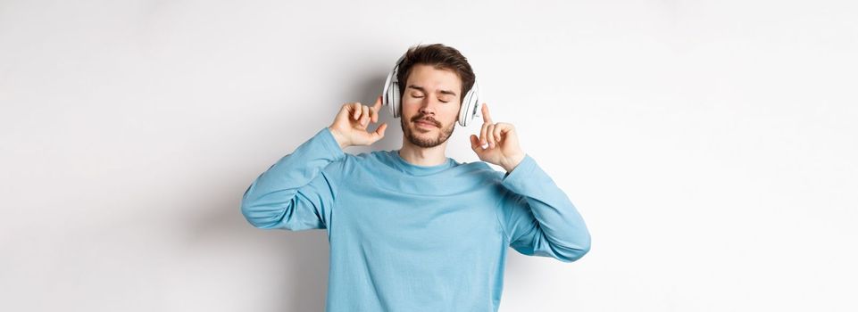 Relaxed young man enjoying favorite song, listening music in headphones with closed eyes and calm face, standing over white background.