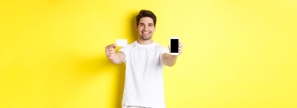 Handsome caucasian male model showing smartphone screen and credit card, concept of mobile banking and online shopping, yellow background.