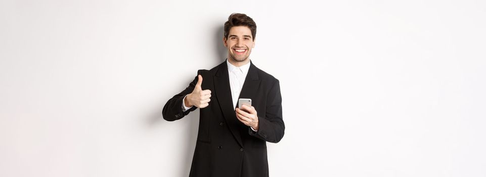 Handsome guy in trendy suit making compliment, showing thumbs-up while using mobile phone, recommending an app or online shop, standing over white background.