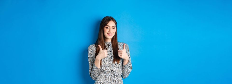 Cheerful smiling woman in dress, showing thumb up and look satisfied, approve and like good thing, recommending product, standing on blue background.