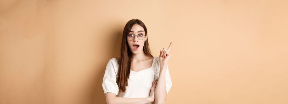 Creative young woman in glasses pitching idea, raising finger in eureka sign and gasping, have a plan, standing on beige background.