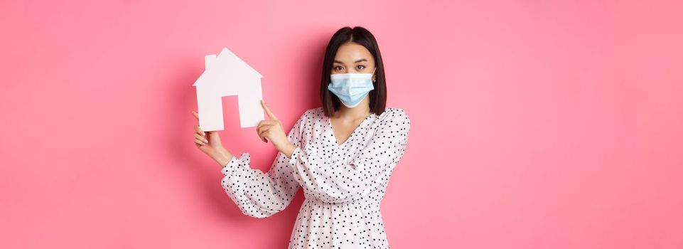 Covid-19, real estate and lifestyle concept. Cute asian woman in face mask selling houses, showing model of home and looking at camera, standing over pink background.