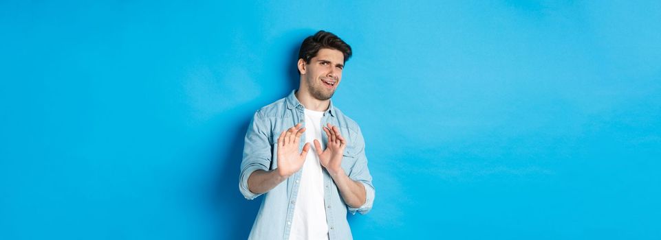 Displeased man refusing, saying no and cringe from something disgusting, standing over blue background.