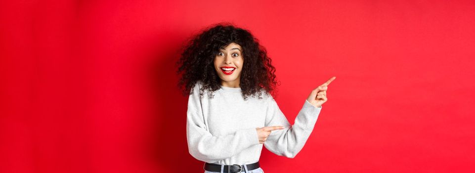 Happy woman with curly hair, smiling amused, pointing fingers left at logo and looking amazed, checking out special deal, standing on red background.