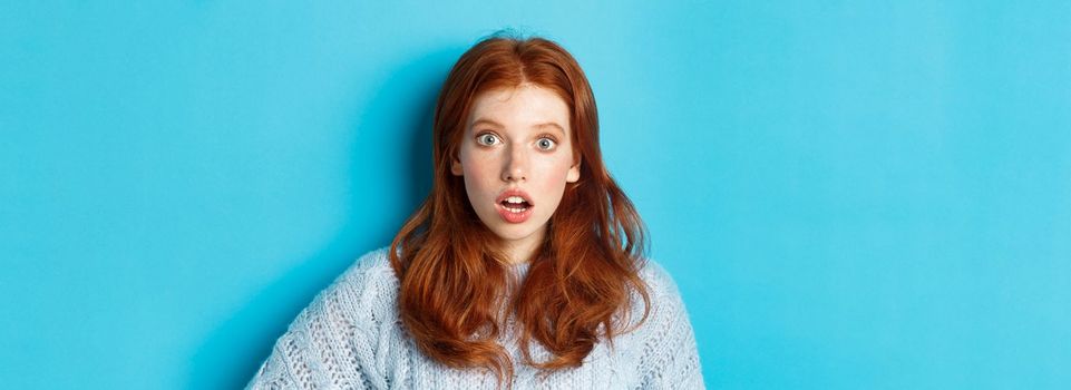 Close-up of startled redhead girl stare at camera speechless, open mouth in awe, standing over blue background.