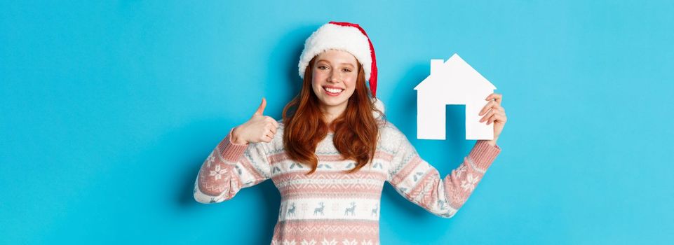 Holiday promos and real estate concept. Satisfied female model with red wavy hair, wearing santa hat and sweater, showing paper house model and thumbs-up, blue background.