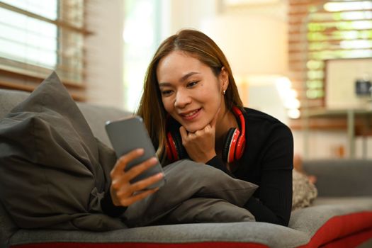 Smiling beautiful woman lying on couch reading chatting with friends via smart phone. Shopping, e-commerce, social media.