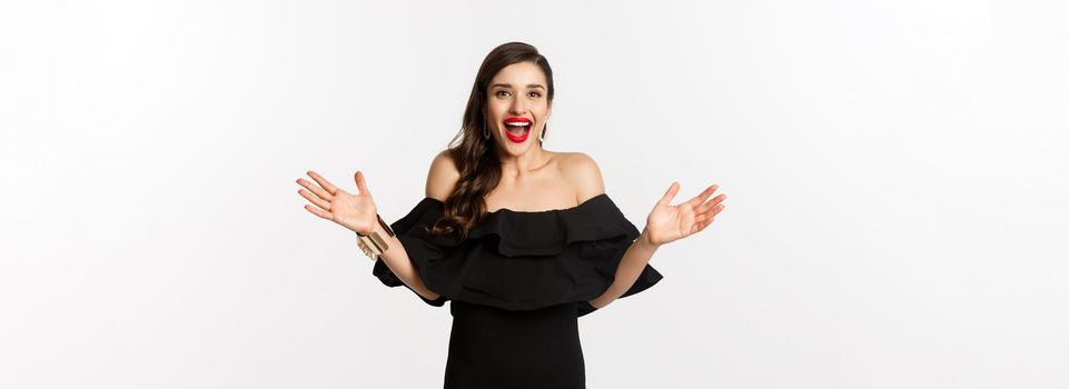 Beauty and fashion concept. Image of surprised and happy young woman in party dress reacting to good news, raising hands up and smiling amazed, white background.