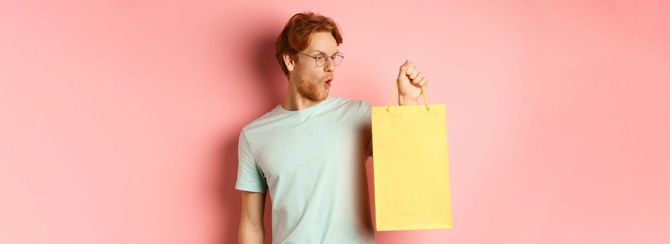 Handsome young man buying presents, holding shopping bag and looking amused, standing over pink background.