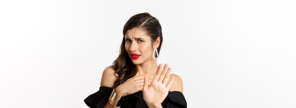 Fashion and beauty concept. Close-up of displeased young woman in black dress saying no, declining offer and looking with disdain, standing over white background.