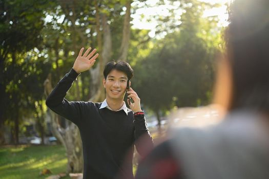 Smiling young asian male walking in campus and greeting his friend with hand raised up.