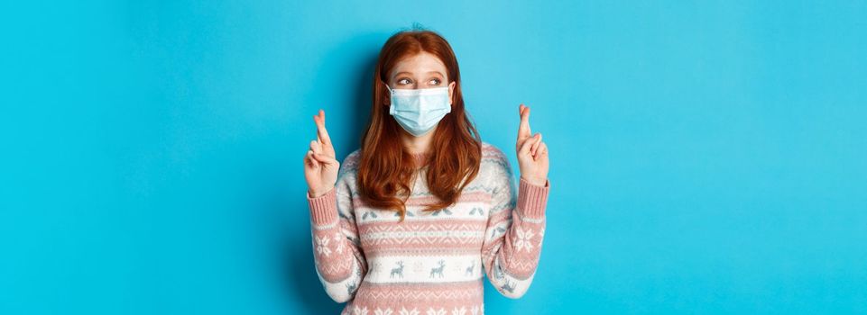 Winter, coronavirus and social distancing concept. Cute hopeful girl with red hair, wearing face mask, cross fingers and making wish, standing over blue background.