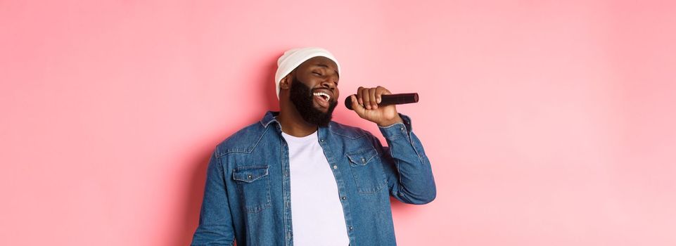 Handsome Black man in beanie and denim shirt singing karaoke, holding microphone, standing over pink background.