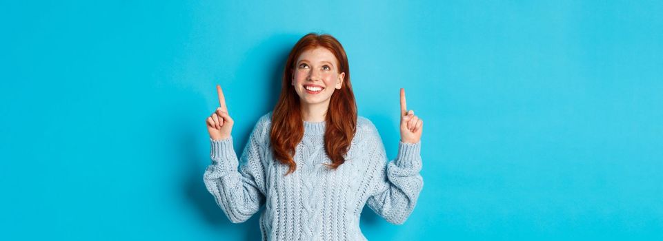 Winter holidays and people concept. Cute teenage redhead girl pointing fingers up, looking at top promo and smiling amused, standing over blue background.