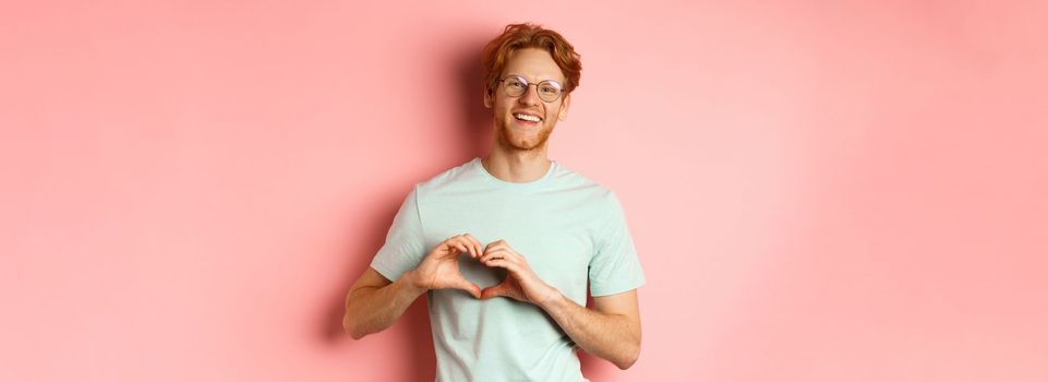 Valentines day concept. Handsome redhead man in glasses, showing heart sign and say I love you, standing over pink background.