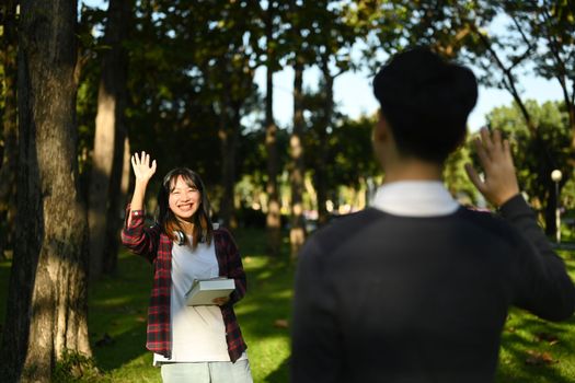 Cheerful asian student woman walking in public park and greeting friend with hand raised up.