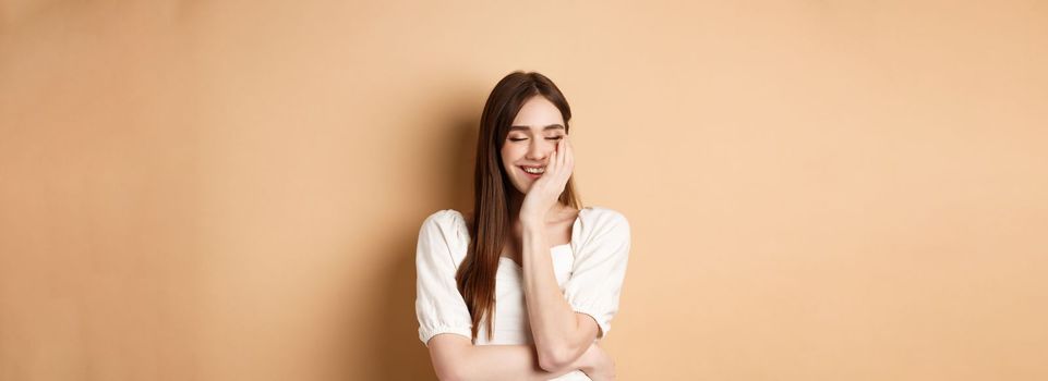 Happy and romantic girl laughing with eyes closed, touching face and feeling joy, standing on beige background.