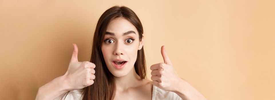 Very cool. Excited girl showing thumbs up and gasping amazed, praising good job, compliment something awesome, standing on beige background.