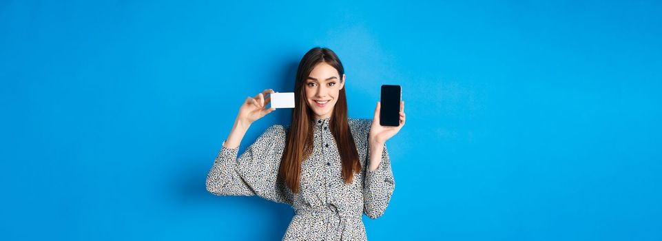 Online shopping. Smiling beautiful girl in dress showing plastic credit card and empty smartphone screen, blue background.