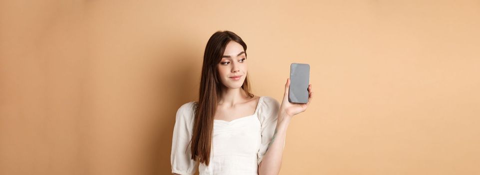 Attractive young woman showing empty smartphone screen, smiling and looking aside, standing on beige background.